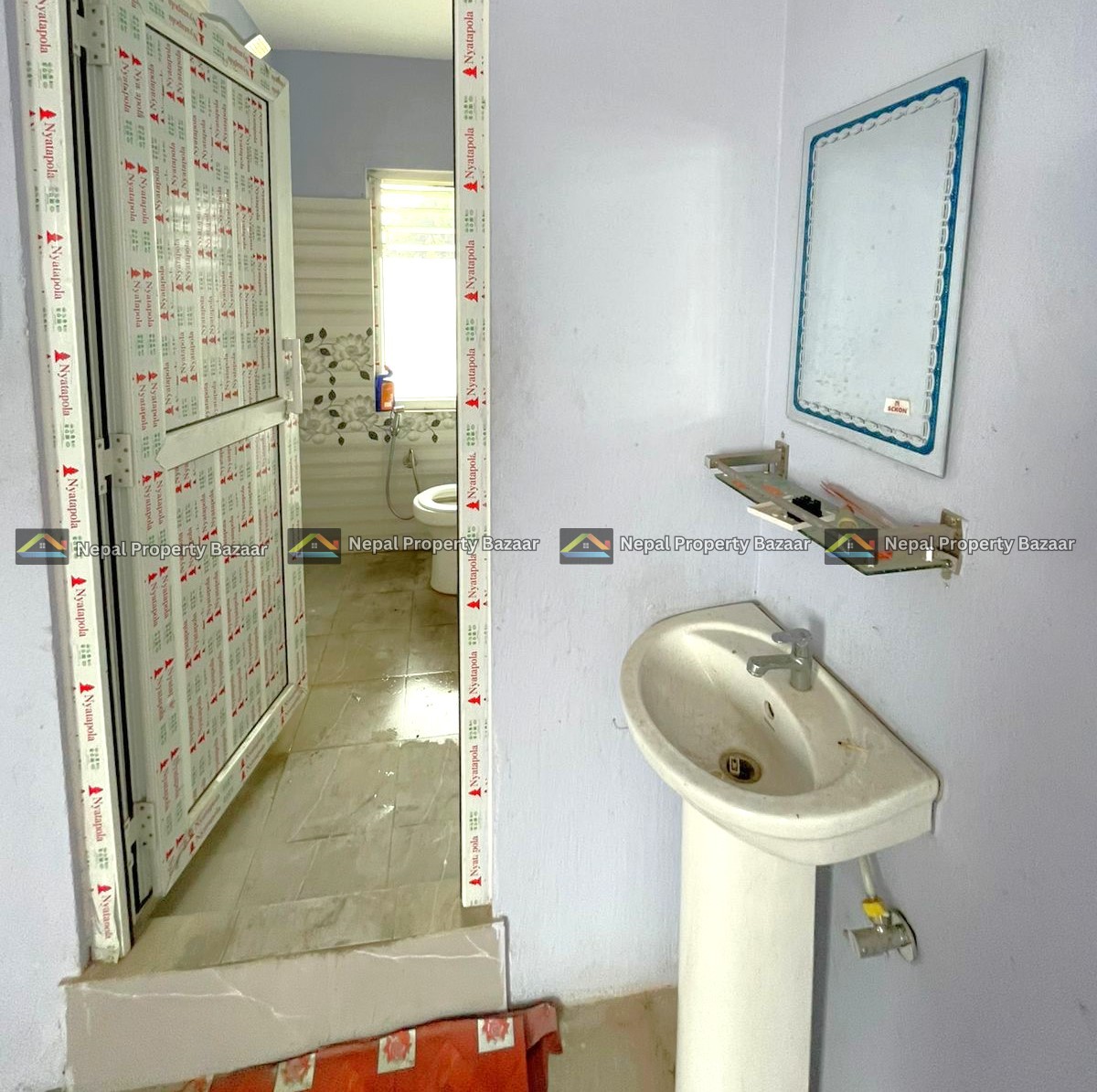 Partially finished bathroom with a pedestal sink, wall mirror, and blue walls; doorway wrapped in protective tape; toilet visible beyond.