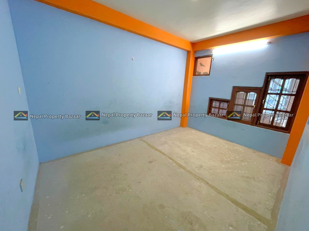 Small empty blue-walled room with orange trim and exposed concrete floor; brown-framed windows with bars along one wall.