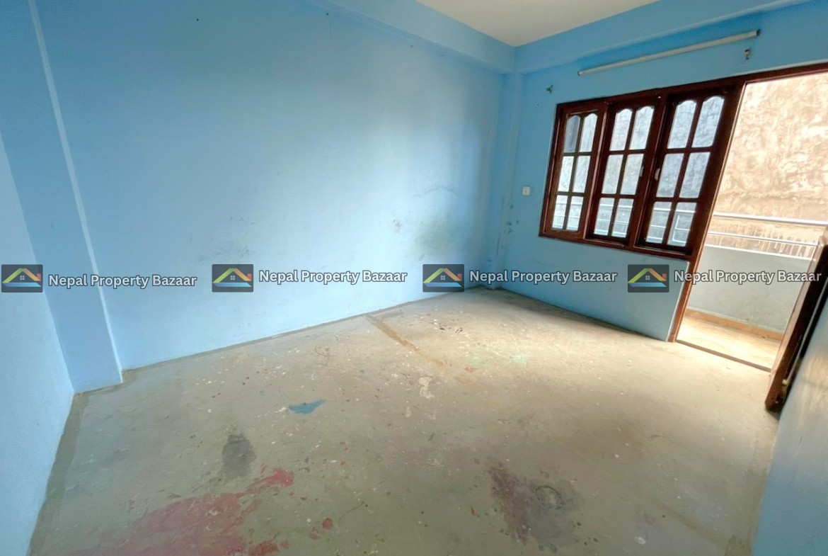 Empty blue-painted room with a concrete floor and paint splatters, featuring large wooden-framed windows that open to a balcony.