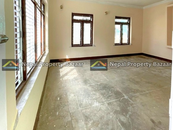 Empty beige-walled room with several wooden-framed windows letting in sunlight; dusty concrete floor.