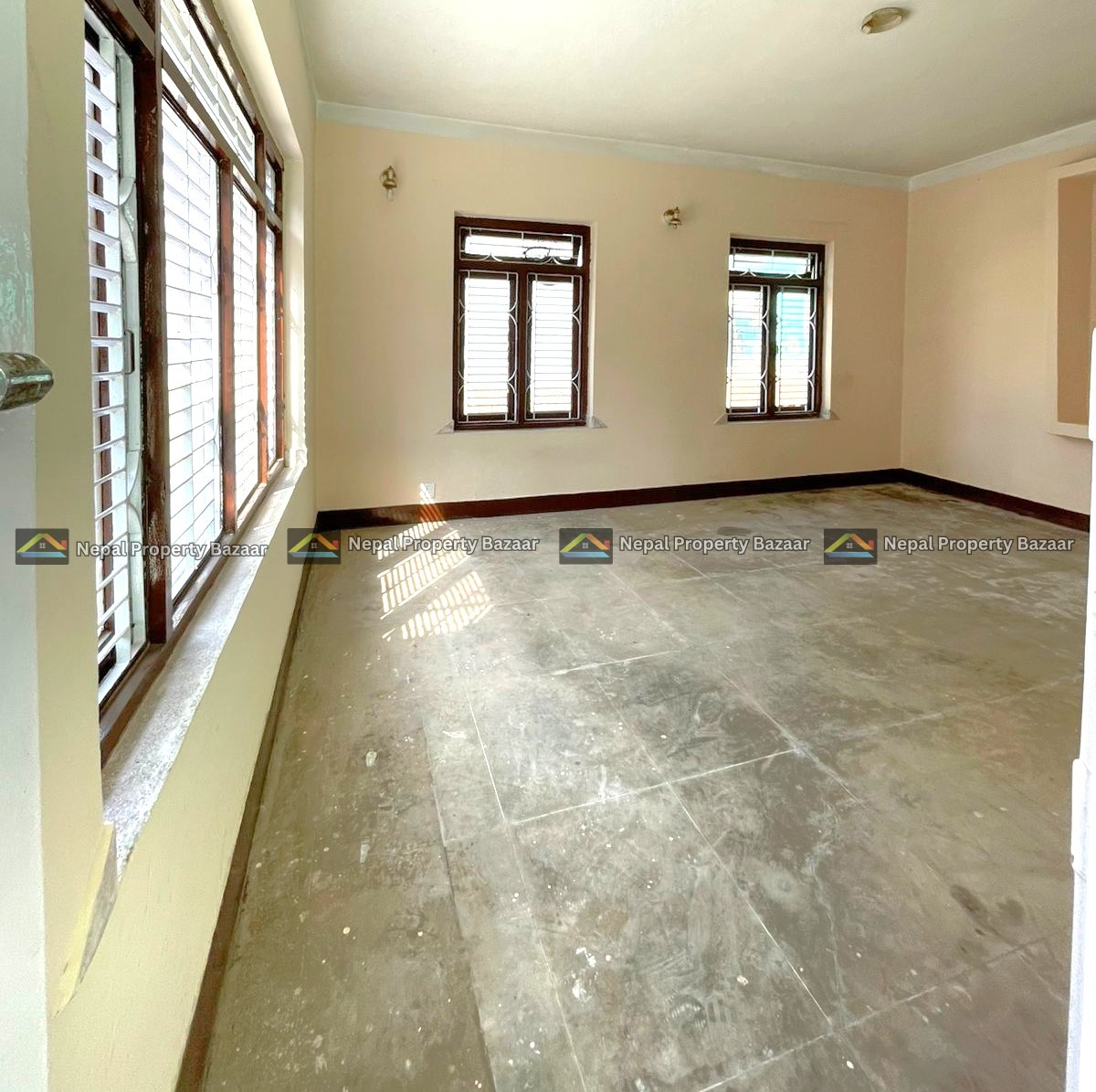 Empty beige-walled room with several wooden-framed windows letting in sunlight; dusty concrete floor.