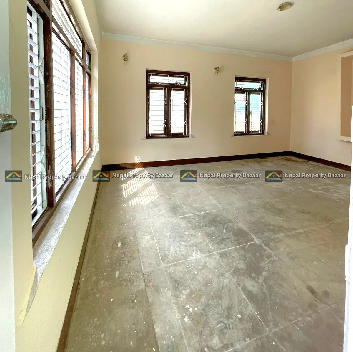 WhatsApp Image 2026-04-19 at 14.17.57 Empty beige-walled room with several wooden-framed windows letting in sunlight; dusty concrete floor.