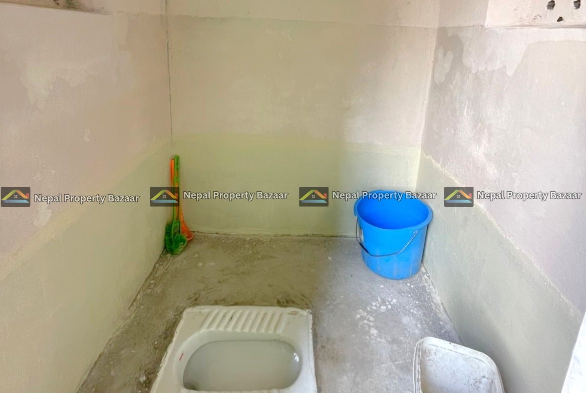 A squat toilet in a bare concrete bathroom; a blue bucket and a white plastic container sit on the floor nearby.