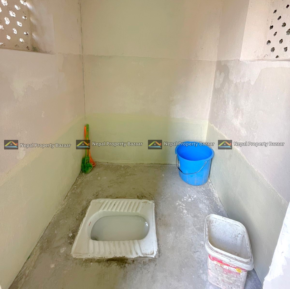 A squat toilet in a bare concrete bathroom; a blue bucket and a white plastic container sit on the floor nearby.